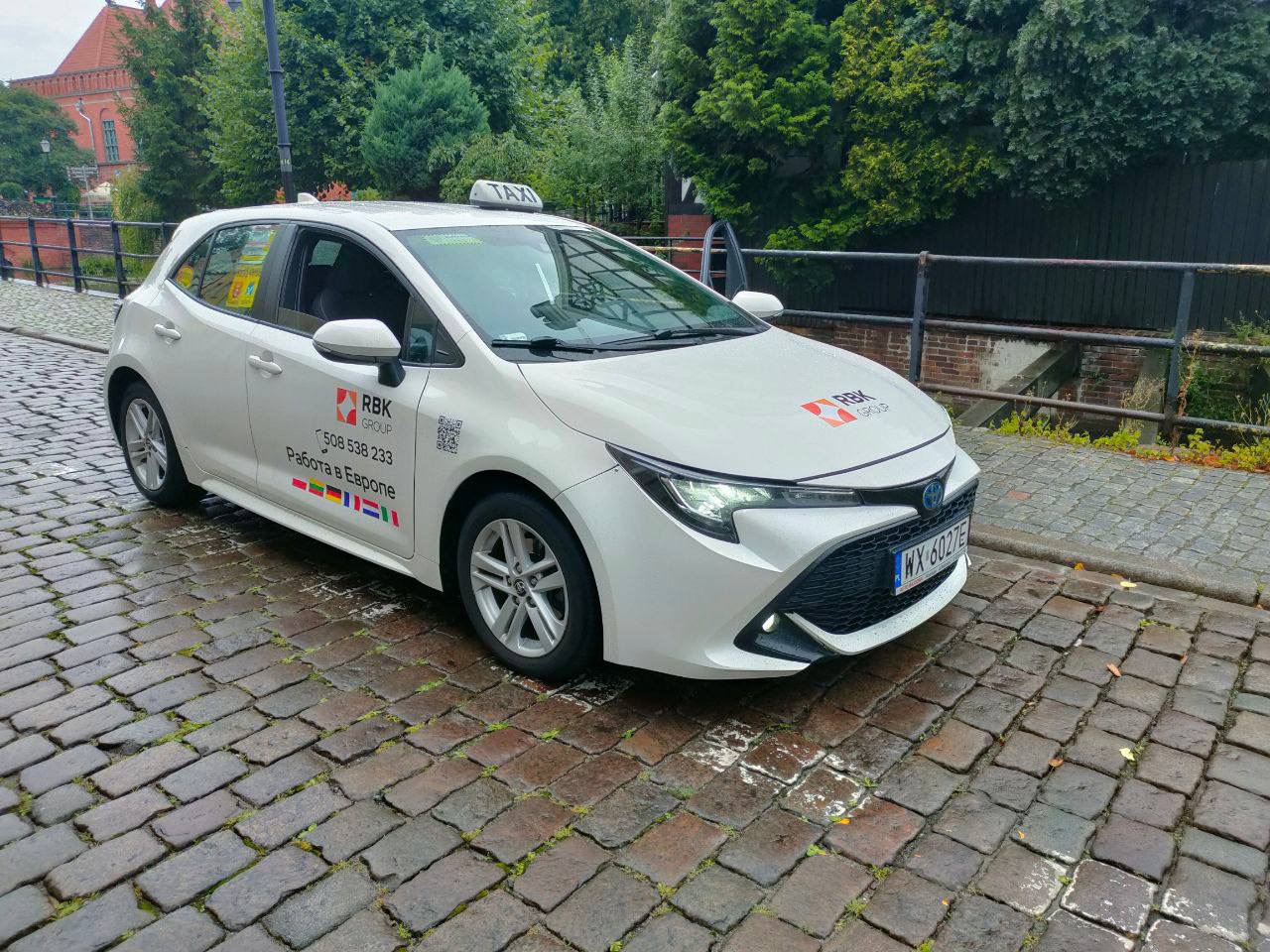 Biała taksówka Toyota Corolla z logo RBK Group na masce i drzwiach, zaparkowana na brukowanej ulicy w pochmurny dzień.