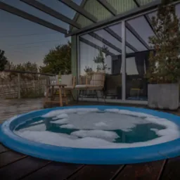 Nastrojowa aranżacja tarasu z okrągłym basenem z bąbelkami, drewnianą podłogą, ażurową altaną z przeszkleniami, rattanowymi fotelami i lampionami.