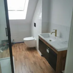 Wykończona łazienka na poddaszu w Kaliszu z kabiną prysznicową z czarnymi ramami, podwieszaną toaletą i umywalką z szafką z drewnianymi akcentami oraz złotą baterią.