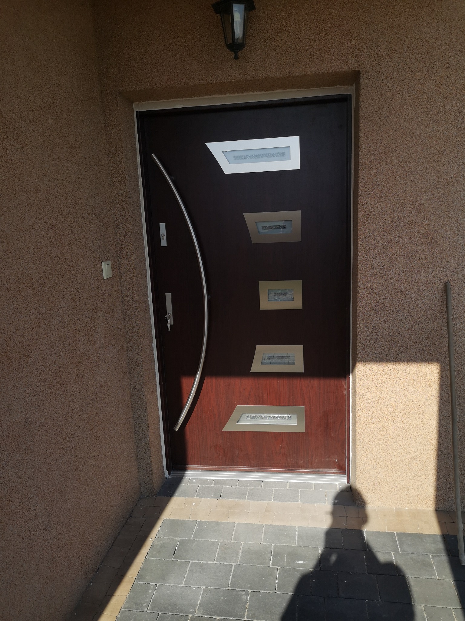 Ciemnobrązowe drzwi wejściowe z czterema prostokątnymi przeszkleniami, chromowaną klamką w kształcie łuku i oświetleniem zewnętrznym, zamontowane w fasadzie o teksturze baranka, z widocznym cieniem...