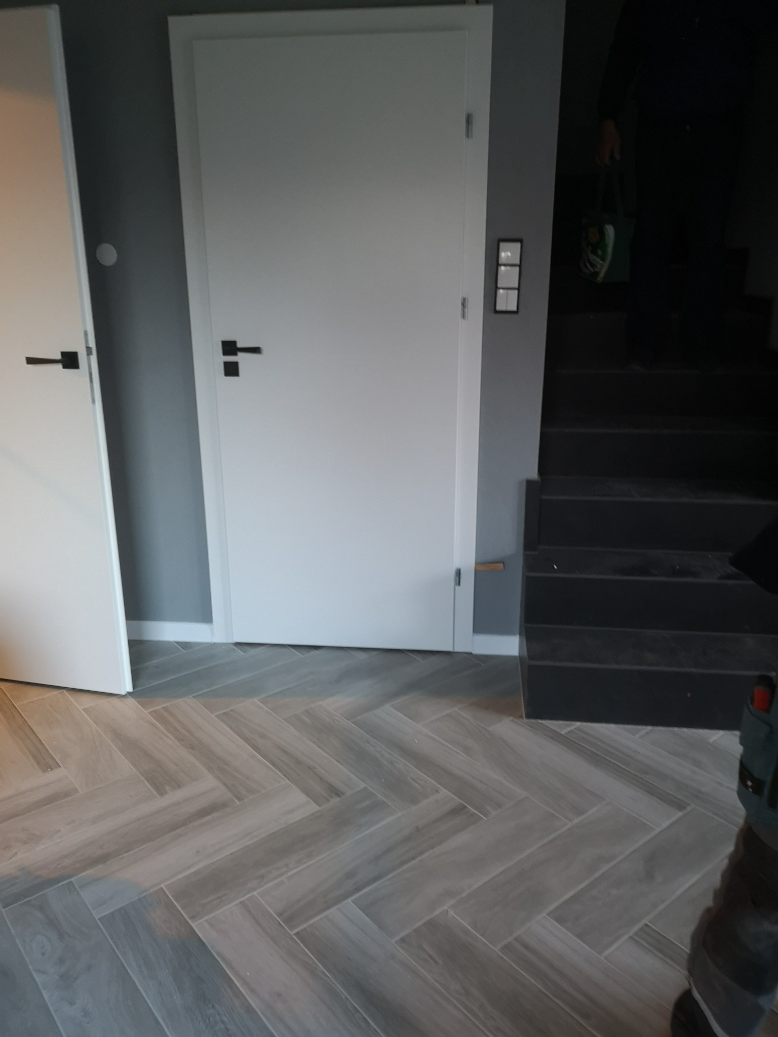 Dwa białe, nowoczesne drzwi z czarnymi klamkami na tle szarej ściany i podłogi wyłożonej szarą parkietową jodełką, widoczne schody w tle.