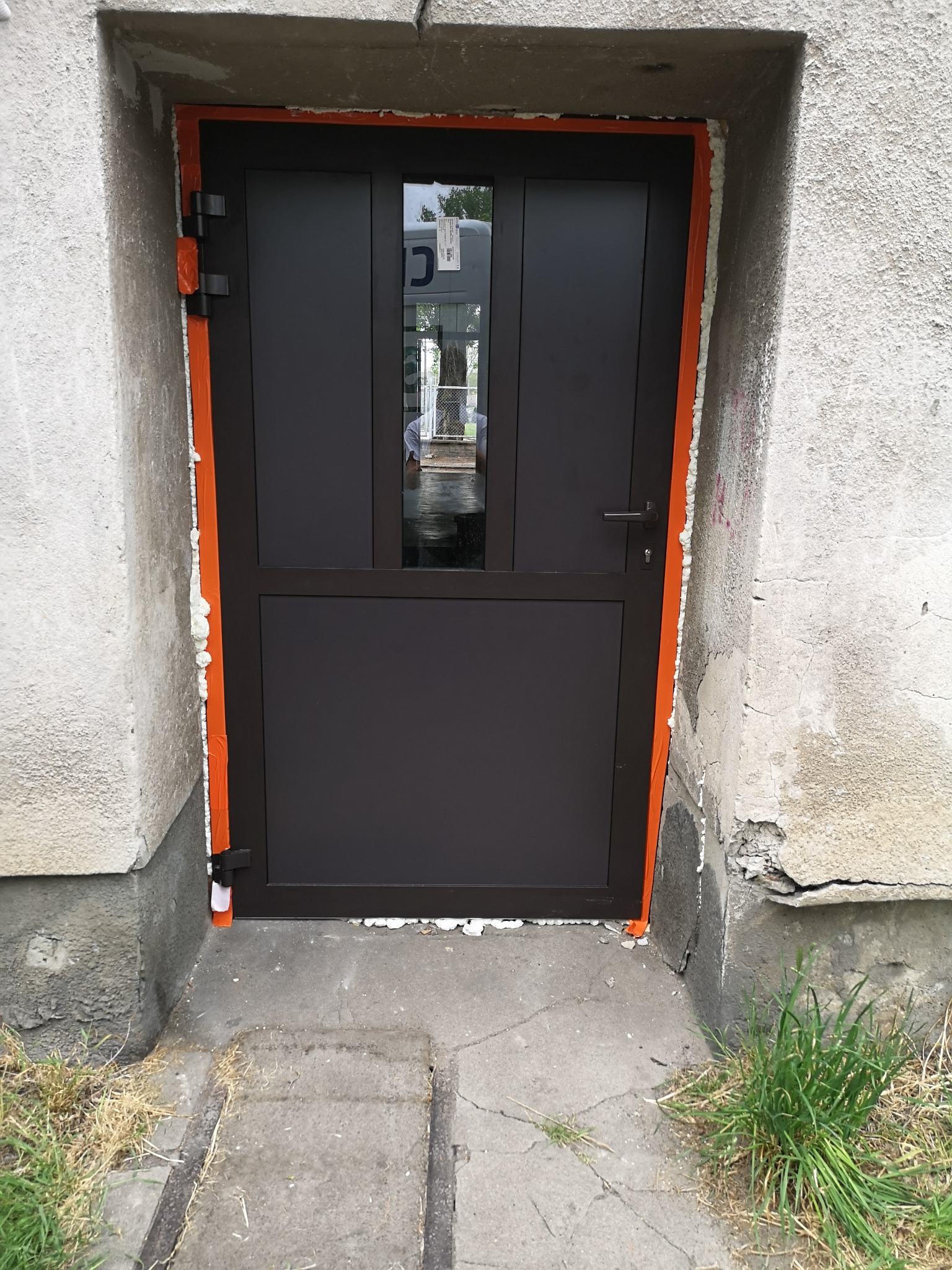 Ciemnobrązowe drzwi wejściowe z szybą, zamontowane w surowym otworze, uszczelnione pianką montażową i pomarańczową taśmą, widoczne ślady na ścianie.