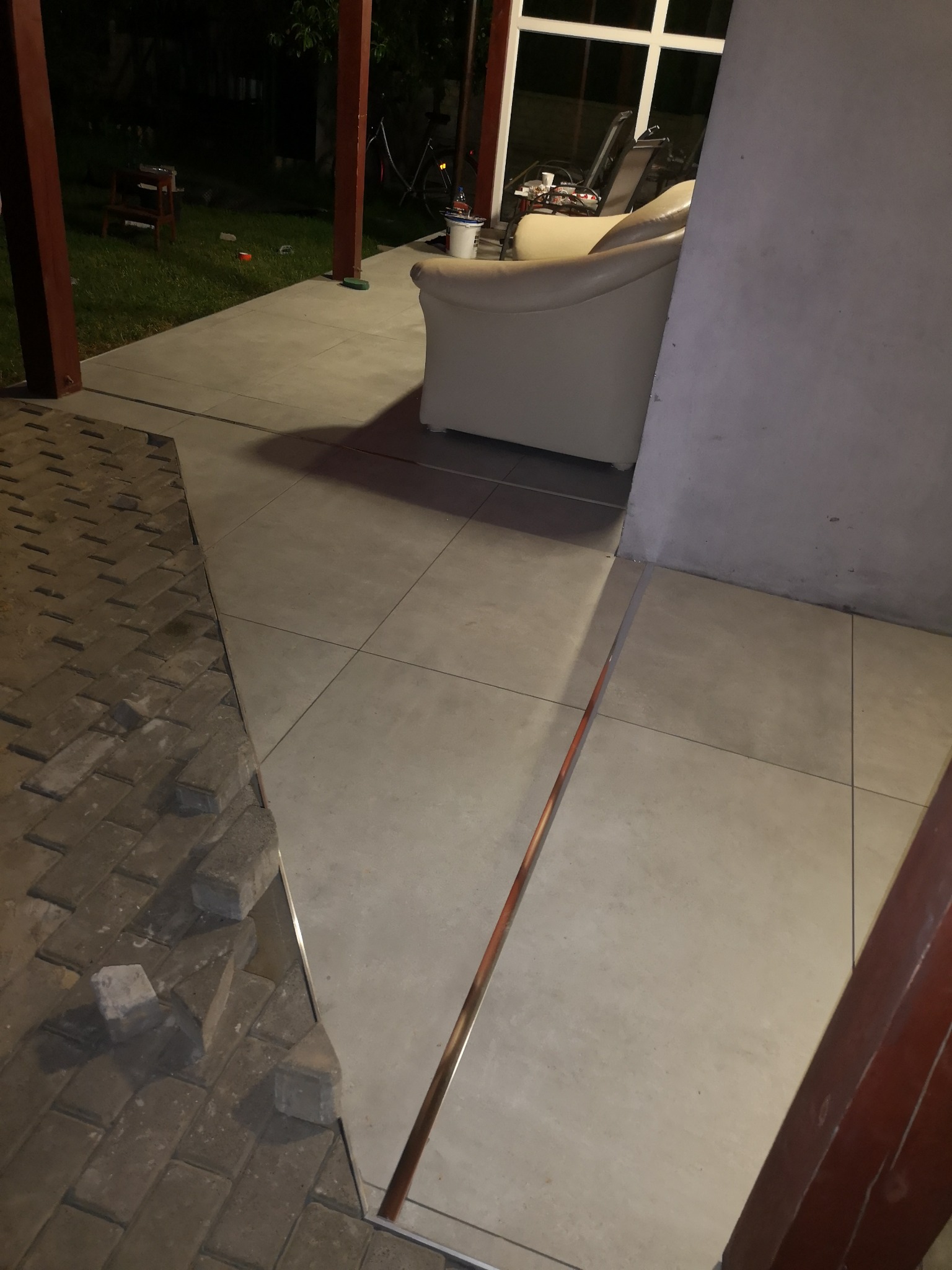 Połączenie nowej, jasnej, kwadratowej podłogi z płytek ceramicznych z kostką brukową na zewnątrz, oddzielone metalową listwą, widok na meble ogrodowe i trawnik w tle.