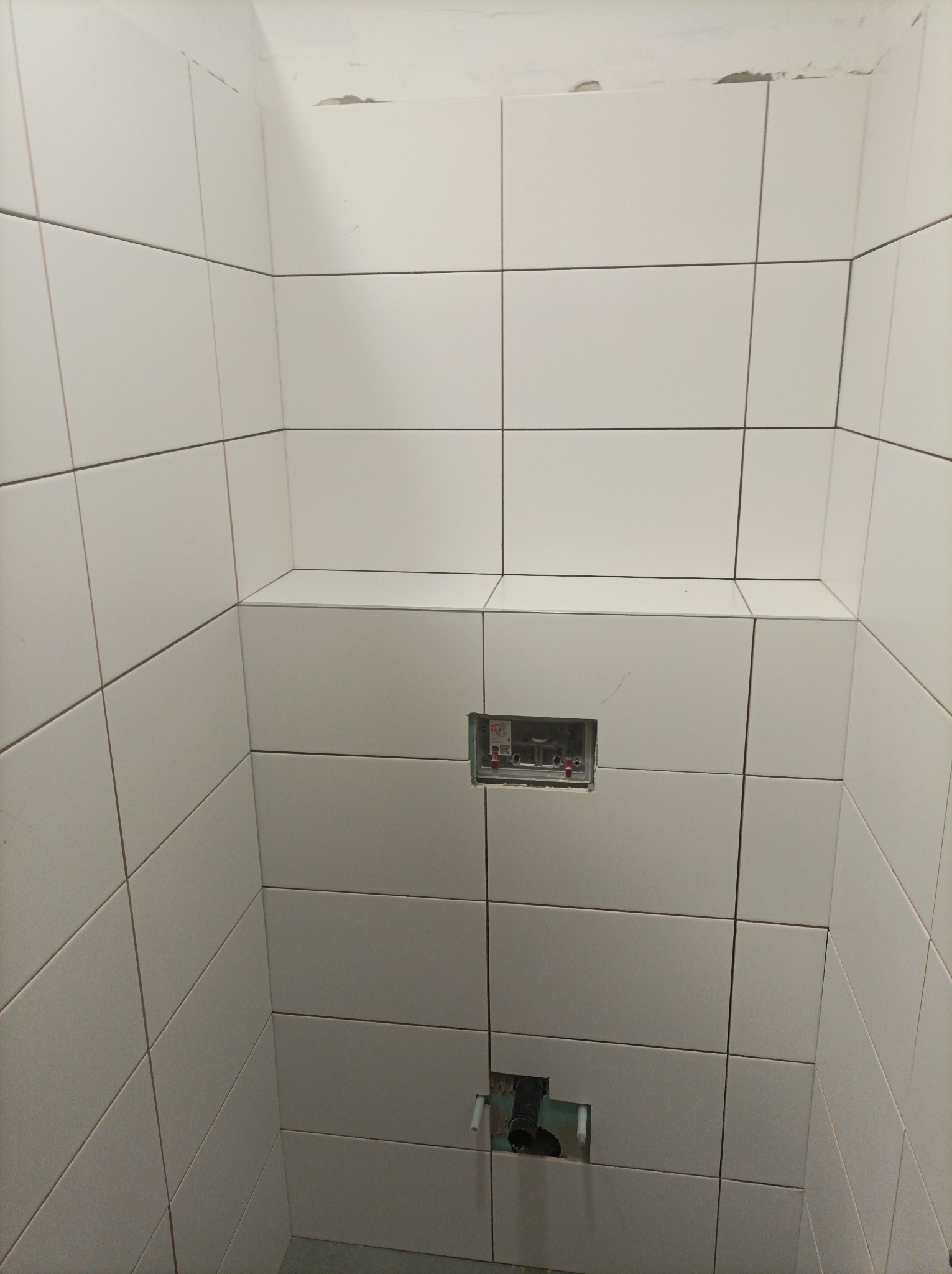 Wnętrze kabiny prysznicowej w trakcie wykańczania, widoczne białe, prostokątne płytki na ścianach, przygotowane otwory na armaturę i niedokończone fugi w górnej części.