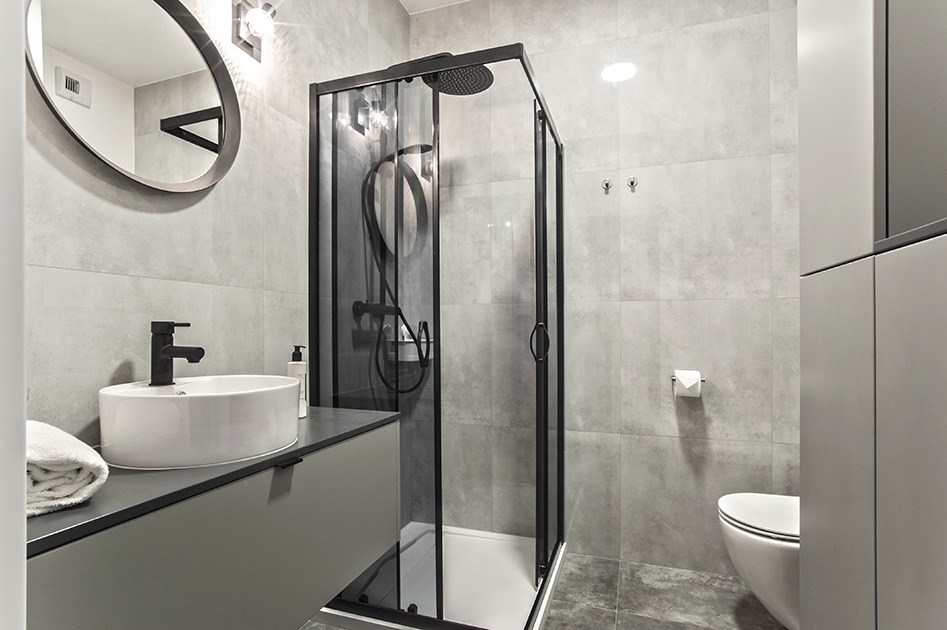 Nowoczesna łazienka w odcieniach szarości z czarnymi akcentami: kabina prysznicowa z czarną ramą, okrągłe lustro z czarną obwódką nad białą umywalką nablatową z czarną armaturą, szare płytki...