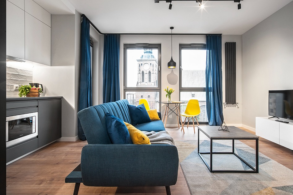 Nowoczesne wnętrze mieszkania z aneksem kuchennym, niebieską sofą z żółtymi poduszkami i widokiem na kościół za oknem.