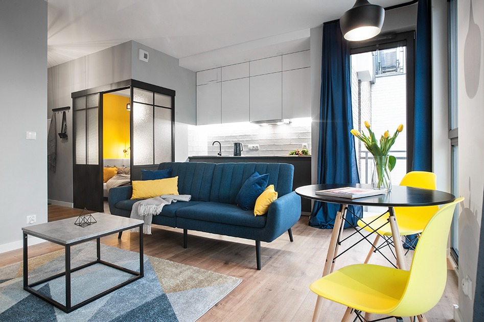 Nowoczesny salon z niebieską sofą, żółtymi akcentami i przeszkloną ścianką działową oddzielającą sypialnię, widok na minimalistyczną kuchnię w tle.