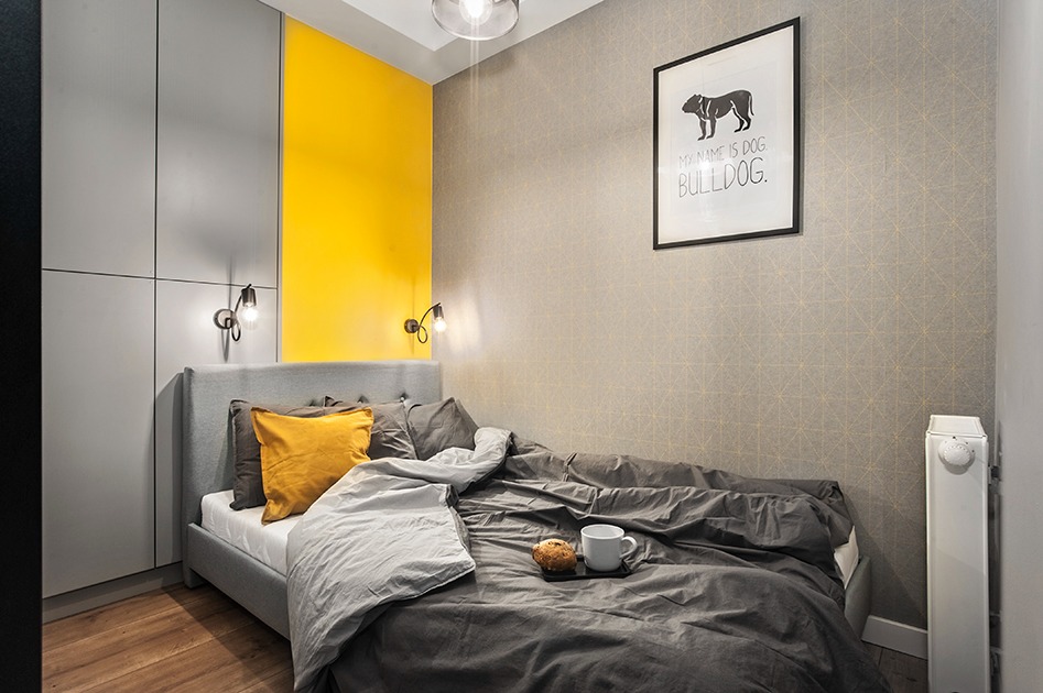 Nowoczesna sypialnia z szarą tapetą w geometryczne wzory, żółtą wstawką na ścianie, szarym łóżkiem z kolorowymi poduszkami, lampkami ściennymi i plakatem z buldogiem.