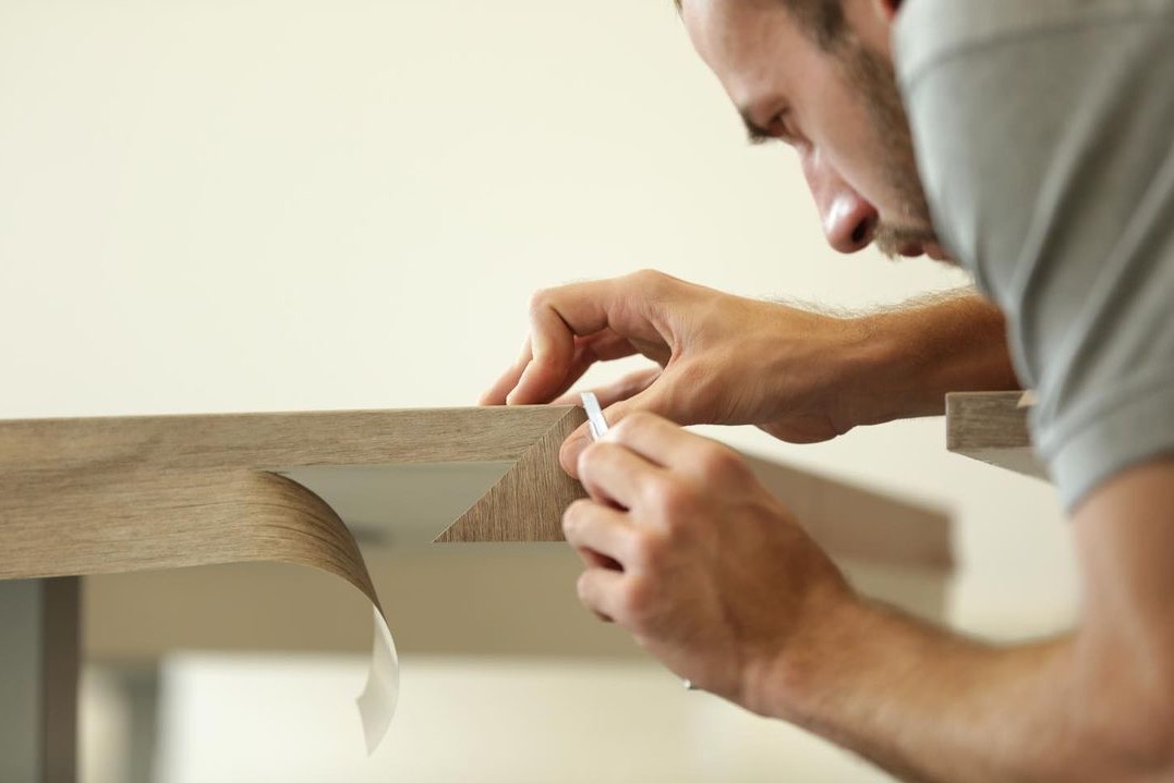 Oklejanie krawędzi blatu meblowego okleiną imitującą drewno, widoczne narzędzie i dłoń pracownika.