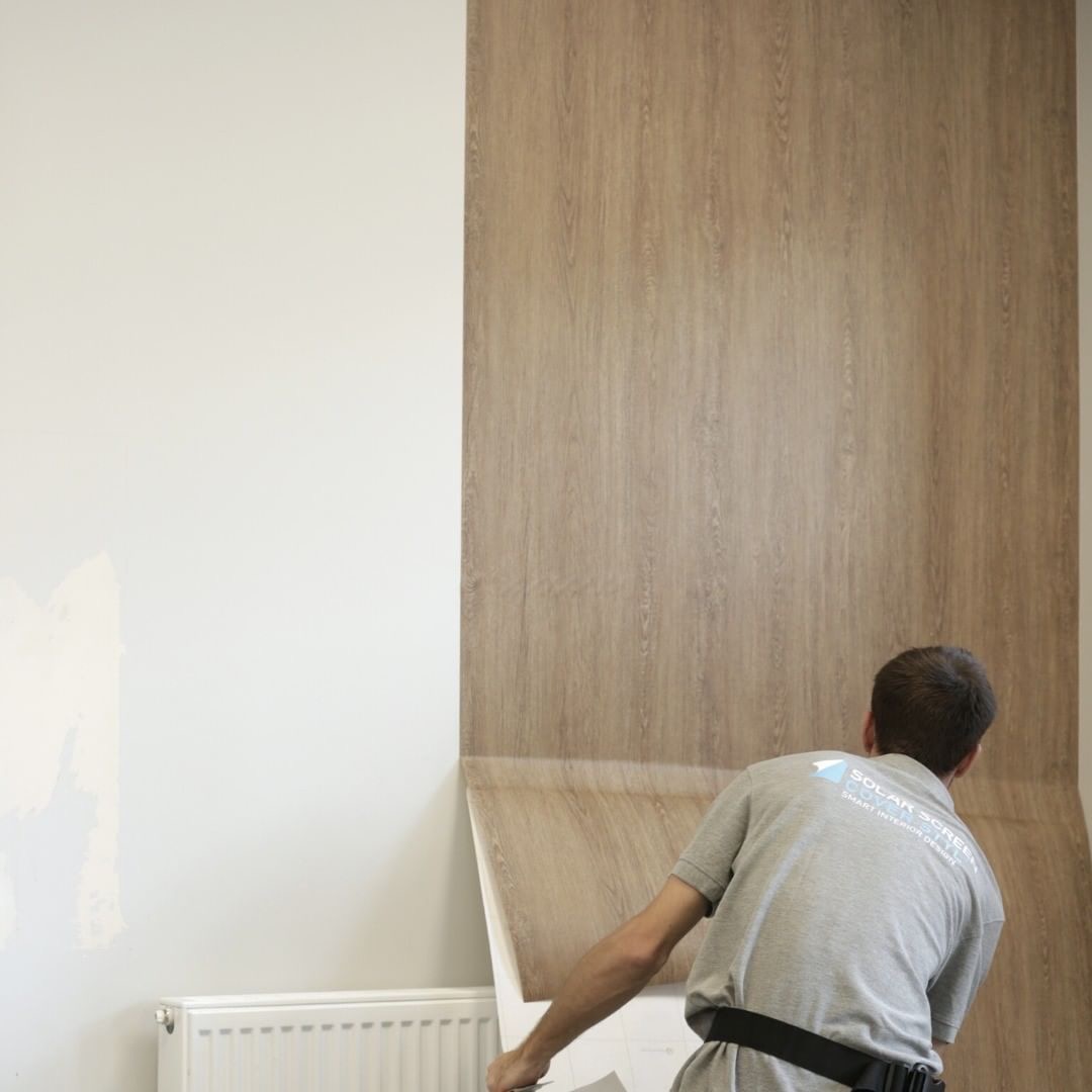Montaż okleiny imitującej drewno na ścianie, widoczny fragment grzejnika i pracownik w szarej koszulce z logo firmy.
