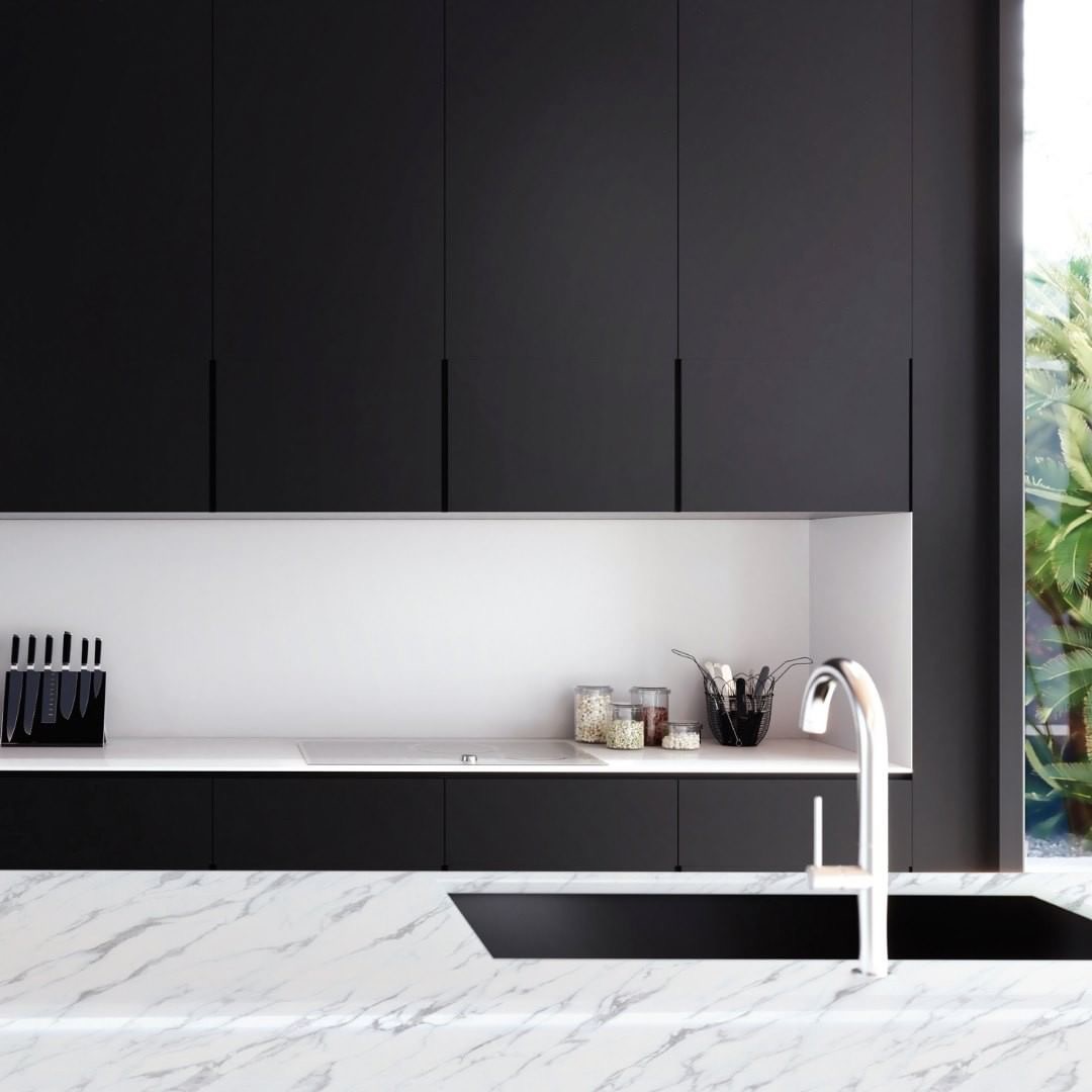 Nowoczesna kuchnia w Krakowie z czarnymi szafkami bez uchwytów, białym blatem i marmurowym wykończeniem wyspy kuchennej, widok na zieleń za oknem.