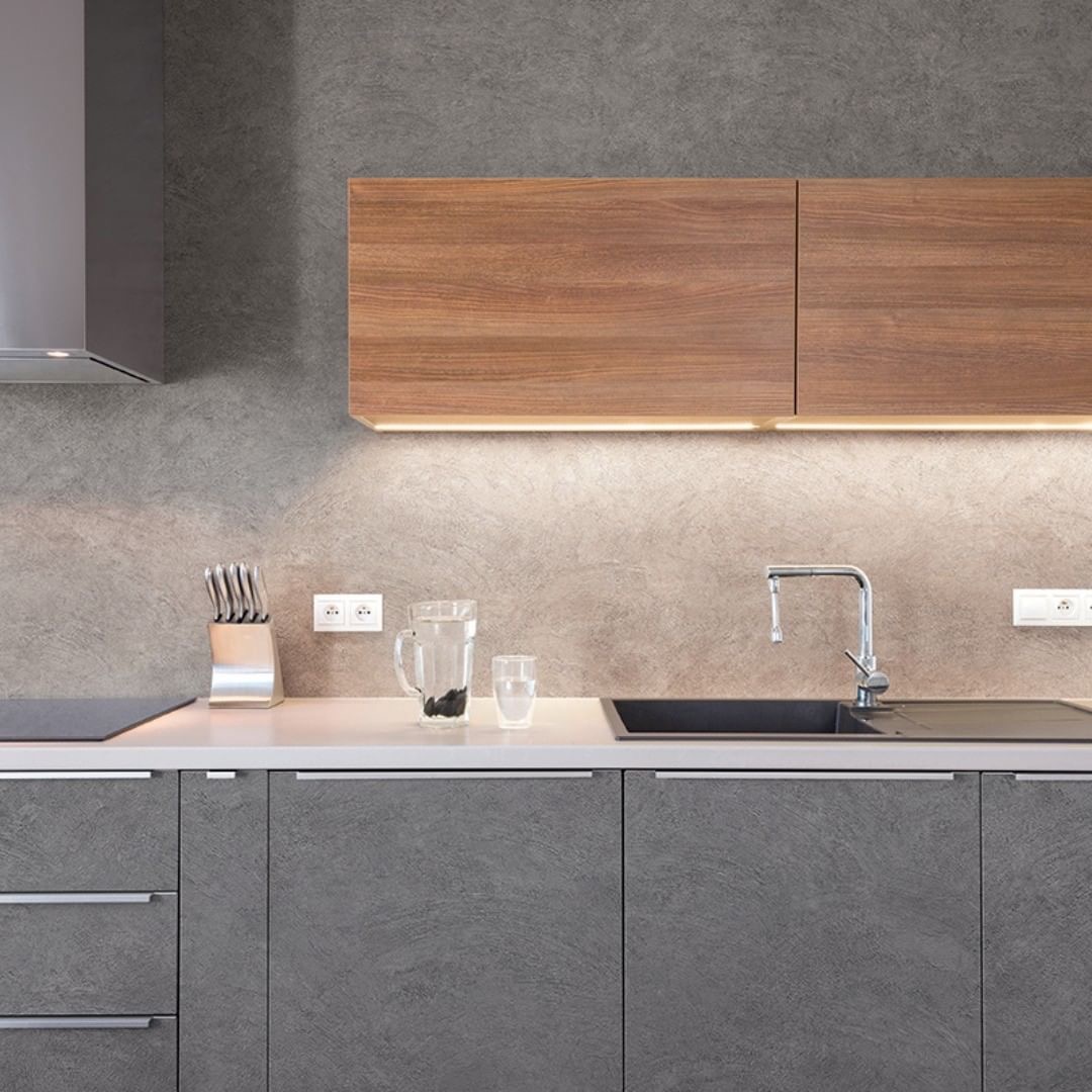 Nowoczesna kuchnia z szarymi frontami szafek o teksturze kamienia, drewnianymi szafkami wiszącymi z podświetleniem LED, srebrnym okapem i białym blatem z czarnym zlewozmywakiem.
