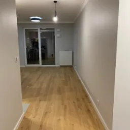 Pusta przestrzeń w nowym mieszkaniu z parkietem, świeżo pomalowanymi szarymi ścianami, grzejnikiem i wyjściem na balkon z barierką zabezpieczającą.