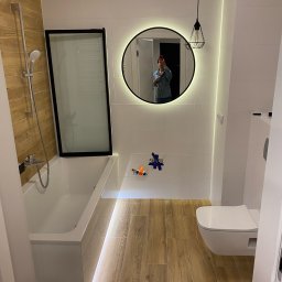 ProEfekt Kompleksowe Wykończenia Wnętrz - Nowoczesna łazienka z drewnopodobną podłogą, białą armaturą, okrągłym lustrem z podświetleniem i lampą wiszącą w stylu industrialnym, widoczne narzędzia na ścianie.