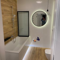 ProEfekt Kompleksowe Wykończenia Wnętrz - Nowoczesna łazienka z drewnopodobnymi płytkami, podświetlaną wanną z czarną szybą, okrągłym lustrem z podświetleniem i wiszącą lampą z metalowym kloszem.