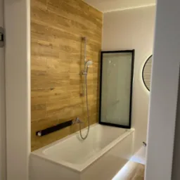 Nowoczesna łazienka z wanną obudowaną białymi płytkami, drewnopodobnymi płytkami na ścianie, kabiną nawannową z czarną ramą oraz oświetleniem LED pod wanną i okrągłym lustrem z podświetleniem.