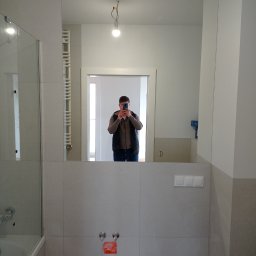 ProEfekt Kompleksowe Wykończenia Wnętrz - Nowoczesna łazienka z dużym lustrem, w którym odbija się osoba robiąca zdjęcie telefonem; widoczna kabina prysznicowa, grzejnik drabinkowy i niedokończone podłączenie baterii.