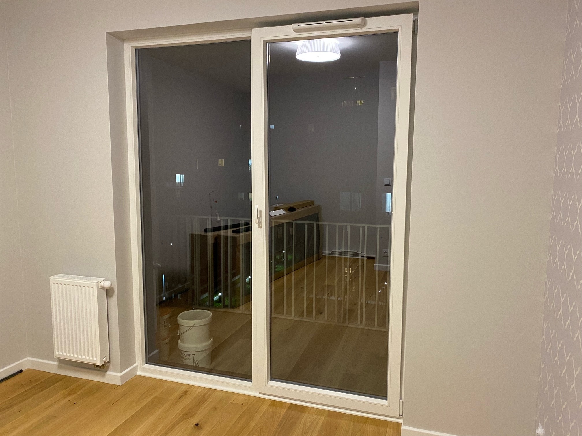 Zamknięte, przesuwne drzwi balkonowe z białymi ramami, widok na balkon z drewnianą podłogą i białą balustradą, grzejnik na ścianie w pokoju.