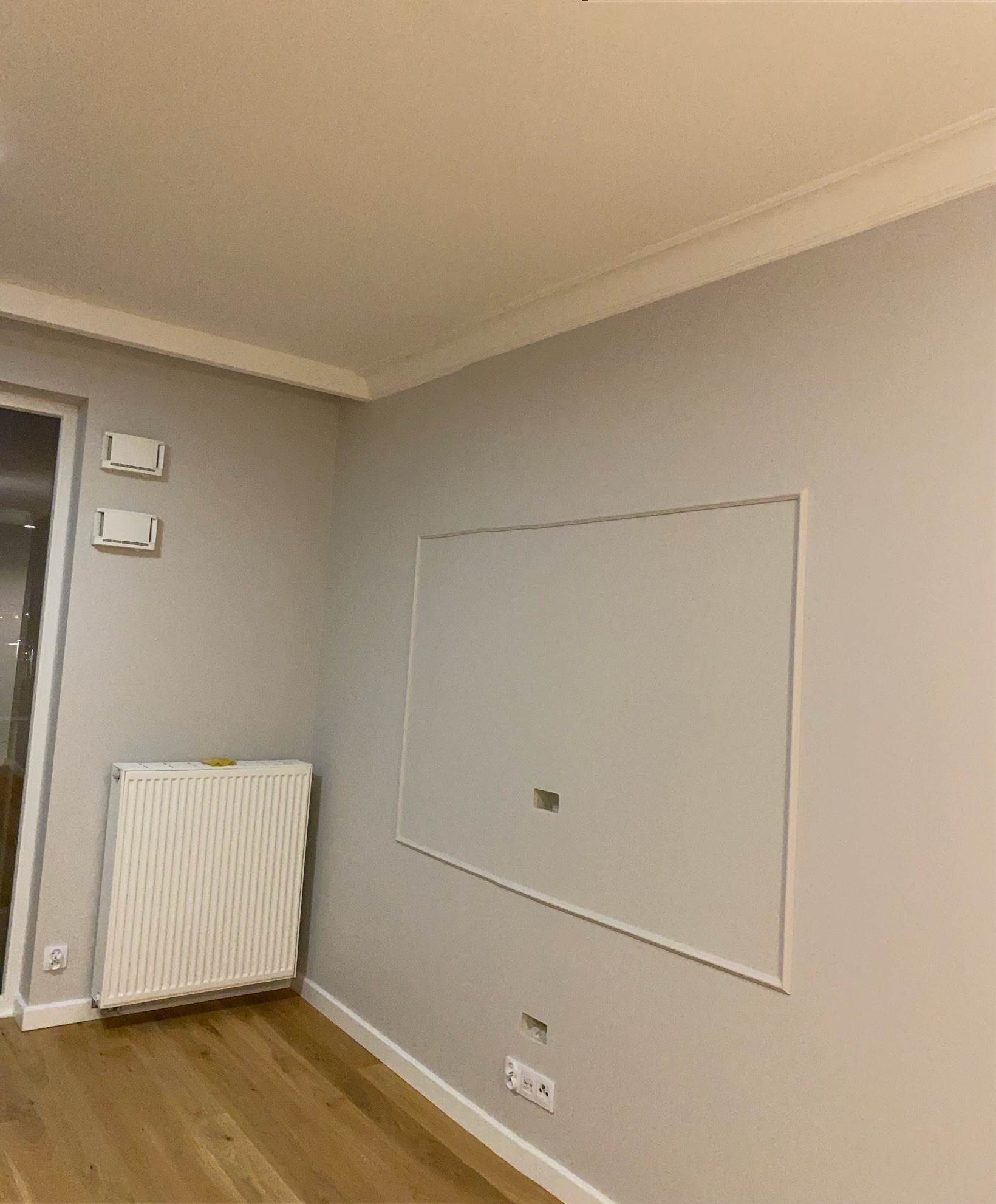 Szare ściany z białymi listwami przypodłogowymi i ramką maskującą otwór rewizyjny, grzejnik oraz parkiet w nowym mieszkaniu w Piastowie po remoncie.