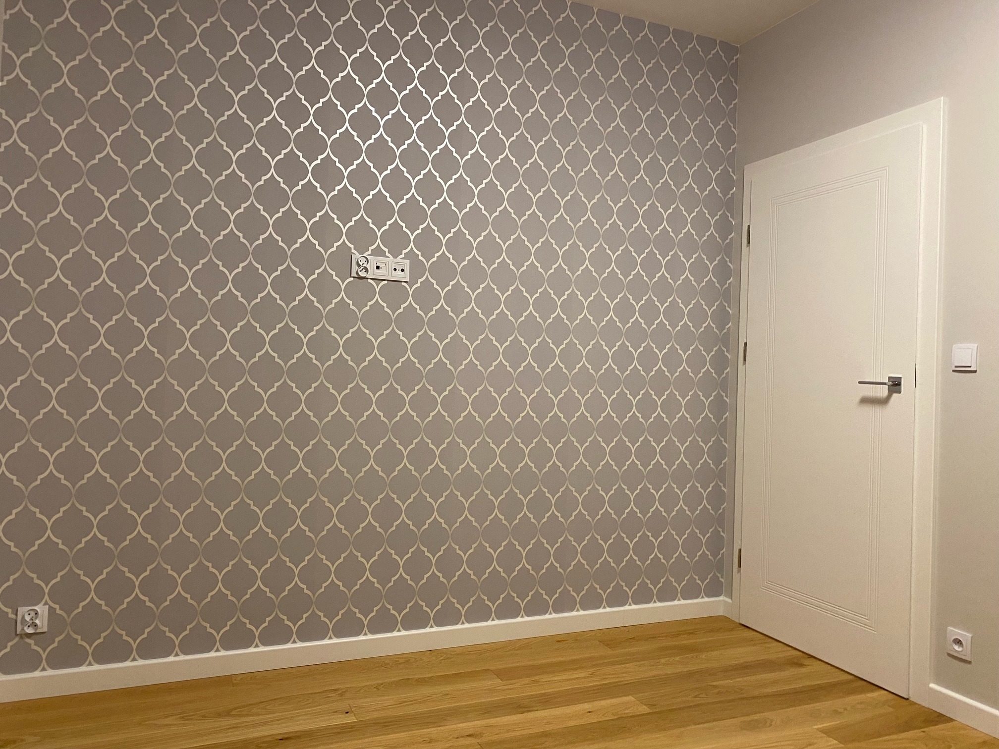 Ściana w pokoju pokryta szarą tapetą z srebrnym marokańskim wzorem, białe listwy przypodłogowe i drewniana podłoga, białe drzwi z srebrną klamką po prawej stronie.
