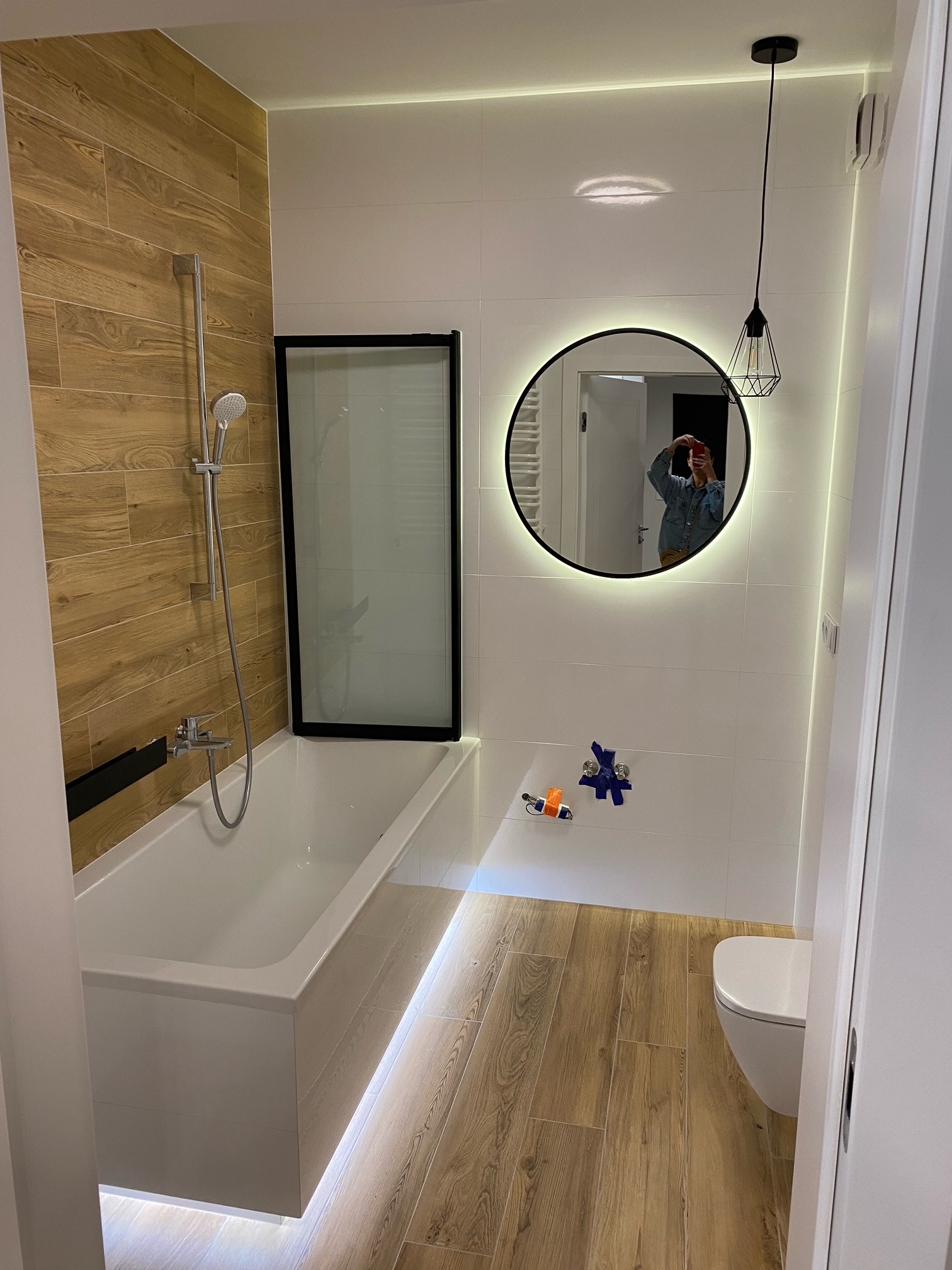 Nowoczesna łazienka z drewnopodobnymi płytkami, podświetlaną wanną z czarną szybą, okrągłym lustrem z podświetleniem i wiszącą lampą z metalowym kloszem.