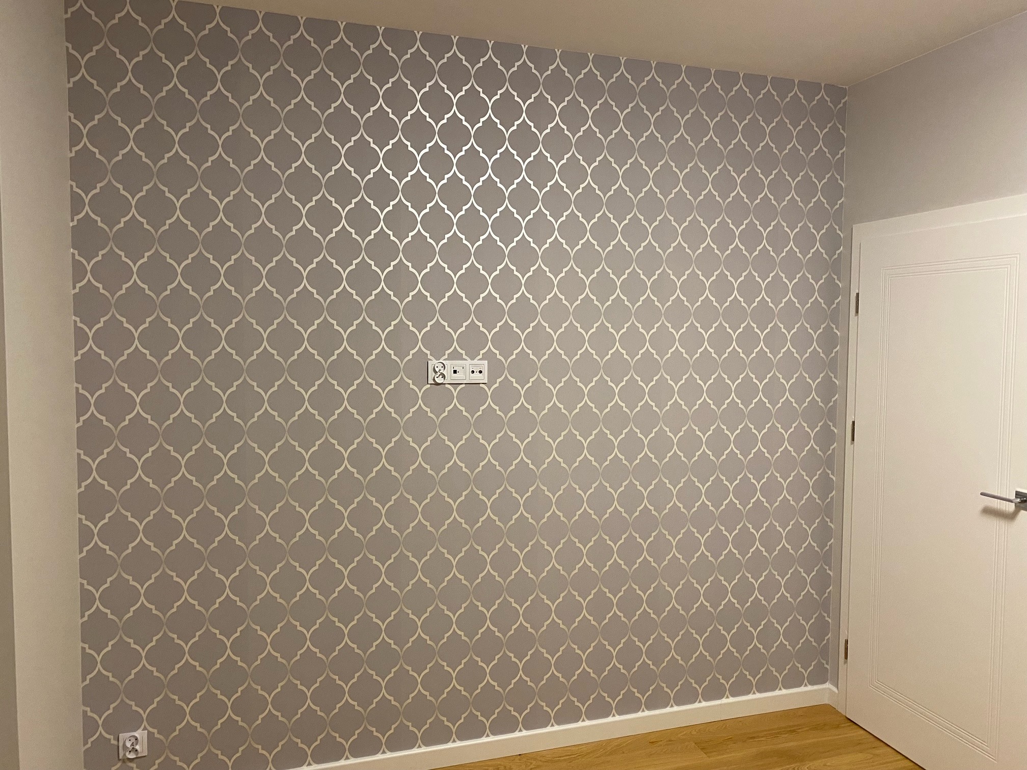 Ściana po tapetowaniu z szarymi tapetami w marokański wzór, widoczne gniazdka elektryczne i fragment białych drzwi.