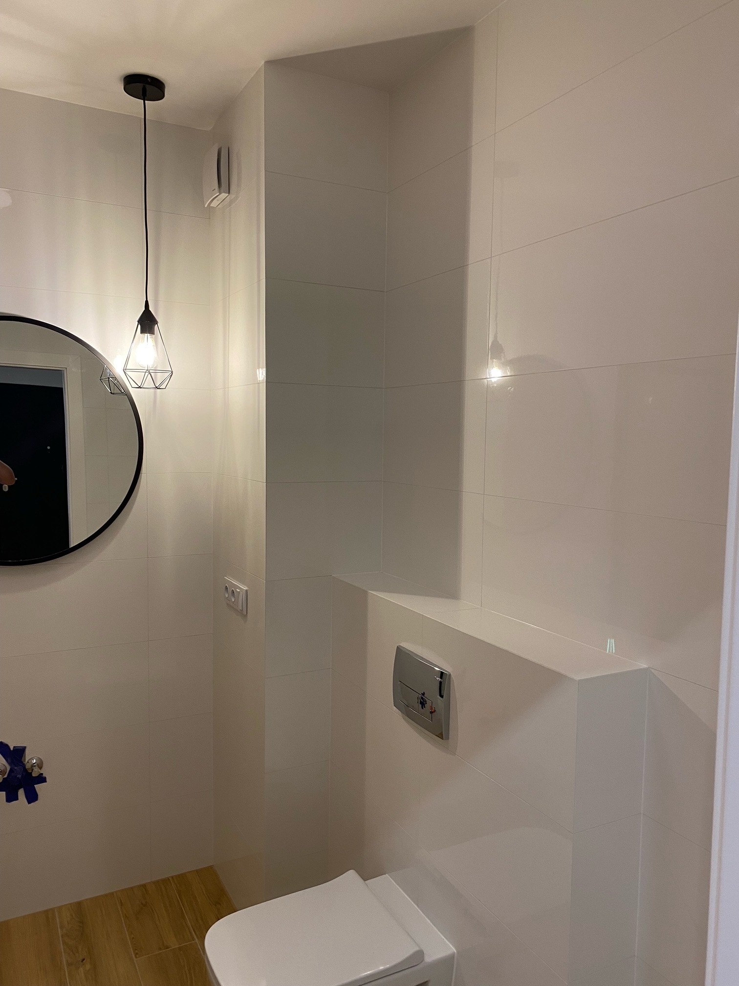 Nowoczesna łazienka z białymi, błyszczącymi płytkami, podwieszaną toaletą, okrągłym lustrem z czarną ramą i designerską lampą wiszącą z geometrycznym kloszem. Na podłodze panele imitujące drewno.