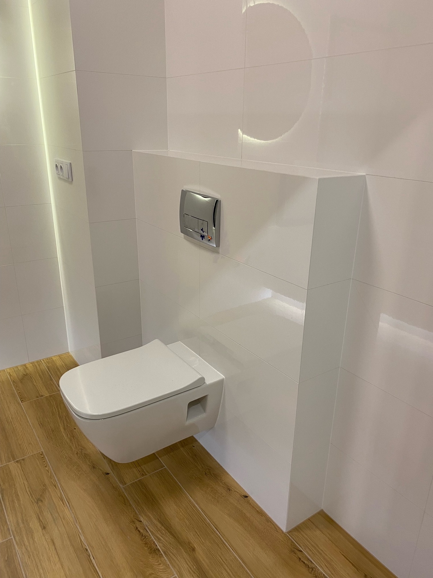 Wisząca toaleta z ukrytym stelażem i chromowanym przyciskiem spłukującym, otoczona białymi, błyszczącymi płytkami, na podłodze drewnopodobna terakota.