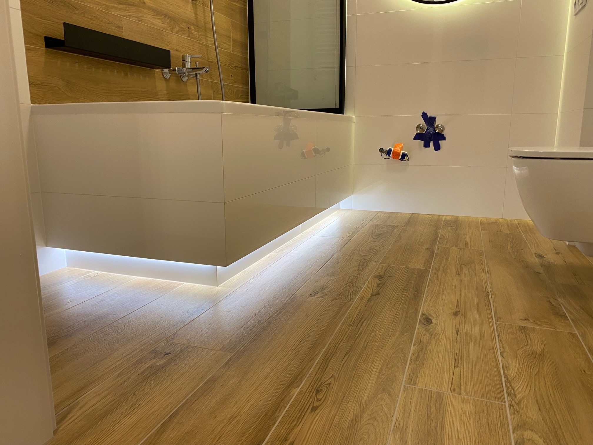 Nowoczesna łazienka z podświetlaną wanną, drewnopodobną podłogą i białymi płytkami ściennymi, widoczne zabezpieczone niebieską taśmą elementy instalacji.