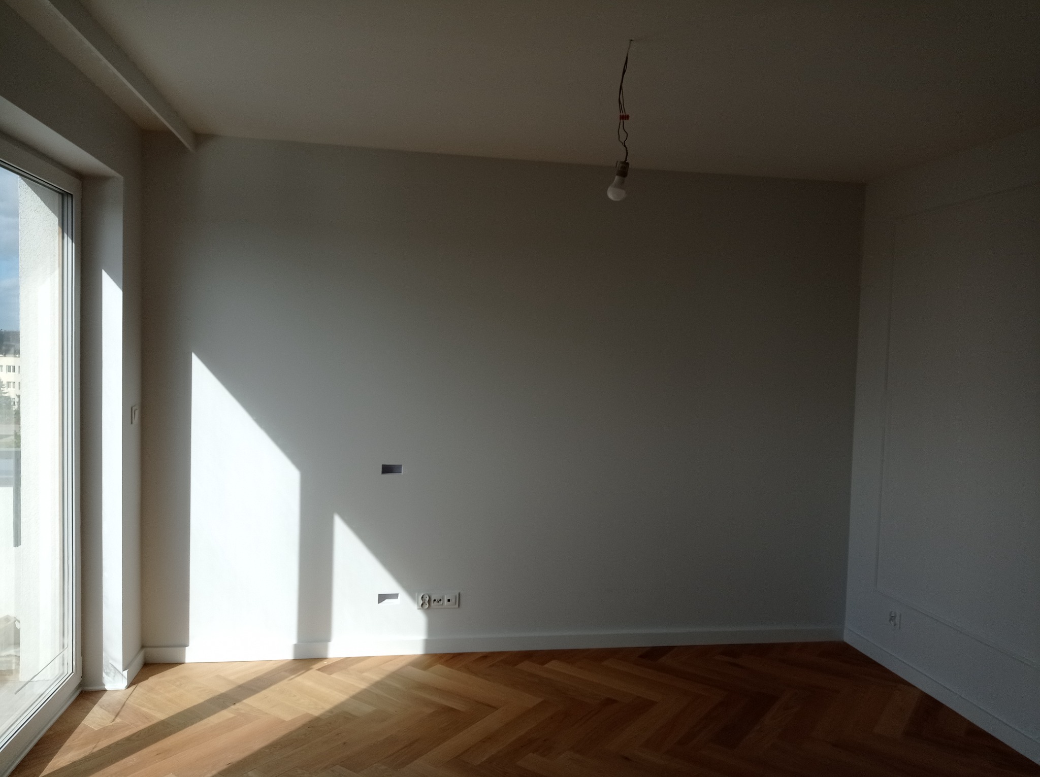 Pusty pokój po malowaniu z parkietem w jodełkę, widoczne gniazdka elektryczne, otwór na instalację i żarówka na kablu.