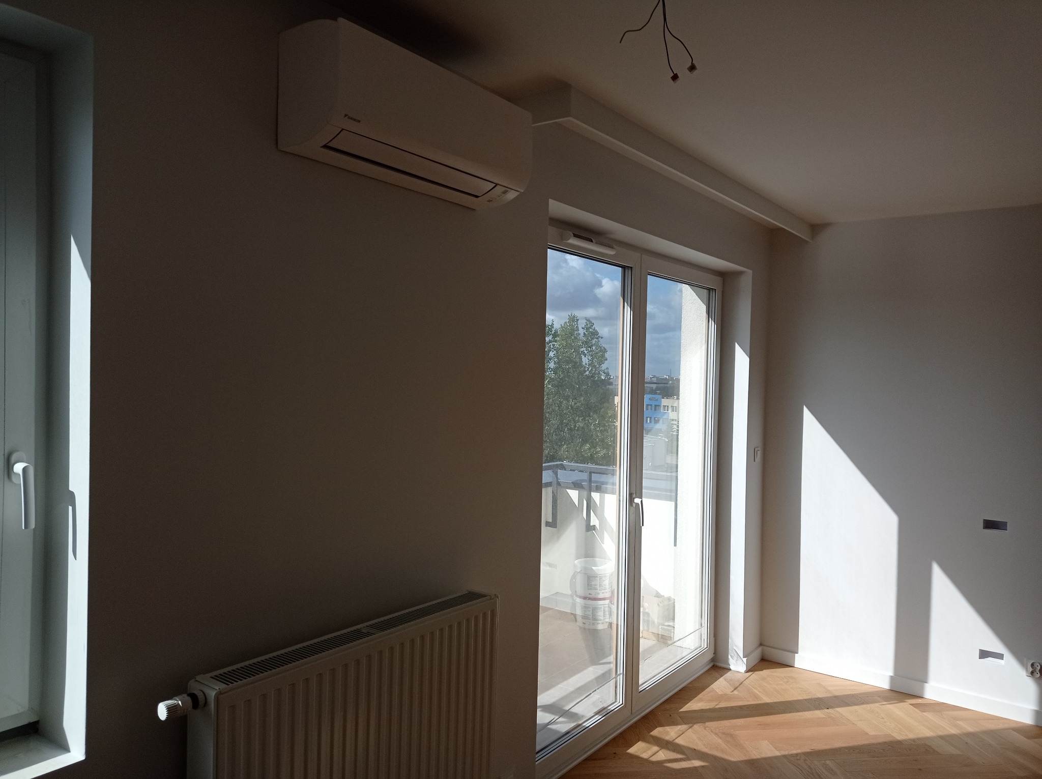 Jasne, odświeżone wnętrze z parkietem w jodełkę, klimatyzatorem Daikin na ścianie i dużym oknem balkonowym z widokiem na zieleń i niebo. Świeżo pomalowane ściany w odcieniach bieli, grzejnik...