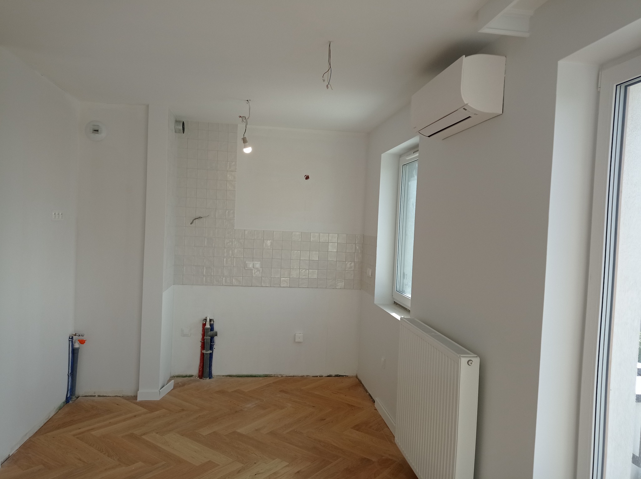 Wnętrze w trakcie prac wykończeniowych: parkiet w jodełkę, częściowo położone płytki na ścianie, grzejnik, klimatyzator, wystające rury instalacji wodnej i elektrycznej, surowe oświetlenie.
