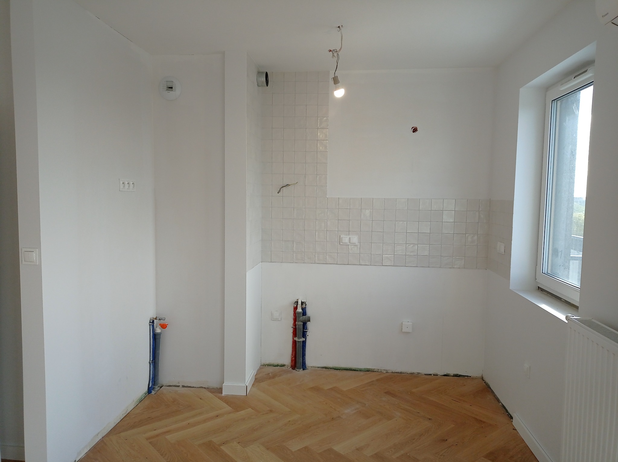 Wnętrze kuchni w trakcie prac wykończeniowych: częściowo ułożone białe kwadratowe płytki na ścianie, parkiet w jodełkę na podłodze, widoczne instalacje wodno-kanalizacyjne, surowy stan wykończenia...