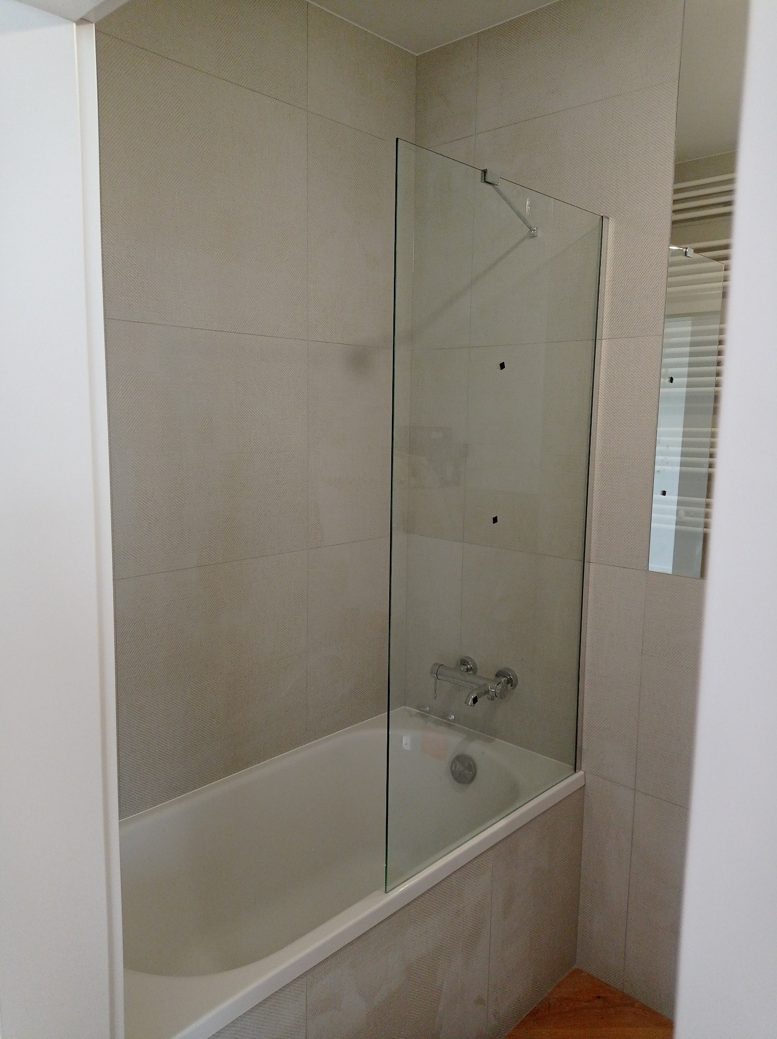 Nowoczesna łazienka z wanną, przeszklonym parawanem nawannowym i beżowymi płytkami o subtelnym wzorze, chromowana bateria. Widoczny fragment grzejnika drabinkowego w odbiciu.