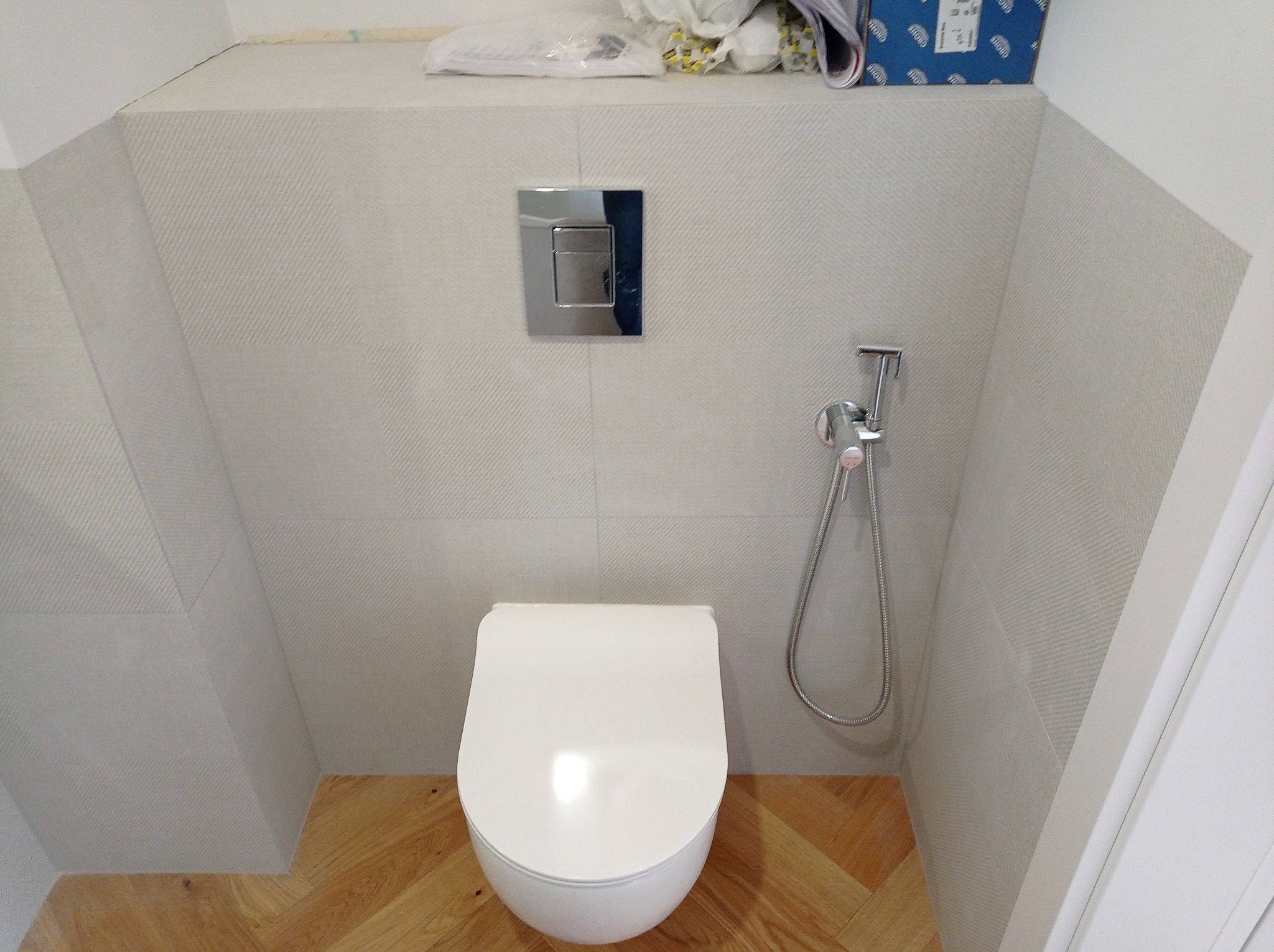 Wisząca toaleta z chromowanym przyciskiem spłukiwania i bidetką na tle jasnych, wzorzystych płytek ceramicznych oraz parkietu ułożonego w jodełkę w wykończonej łazience.