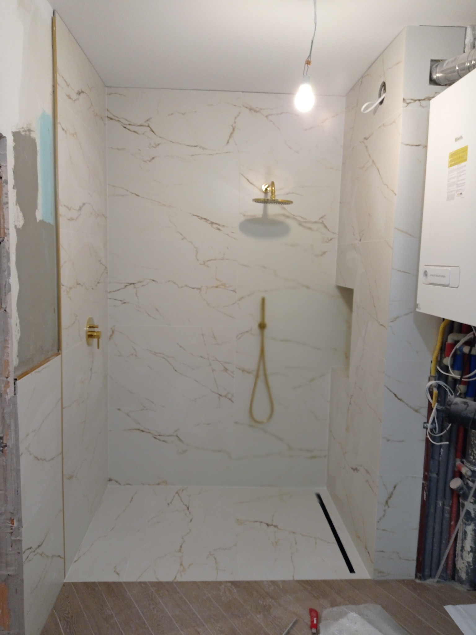 Nowoczesna kabina prysznicowa z marmurowymi płytkami w odcieniach bieli i złota, z zamontowaną deszczownicą i baterią w kolorze złotym, widoczne fragmenty instalacji wodnej i grzewczej...