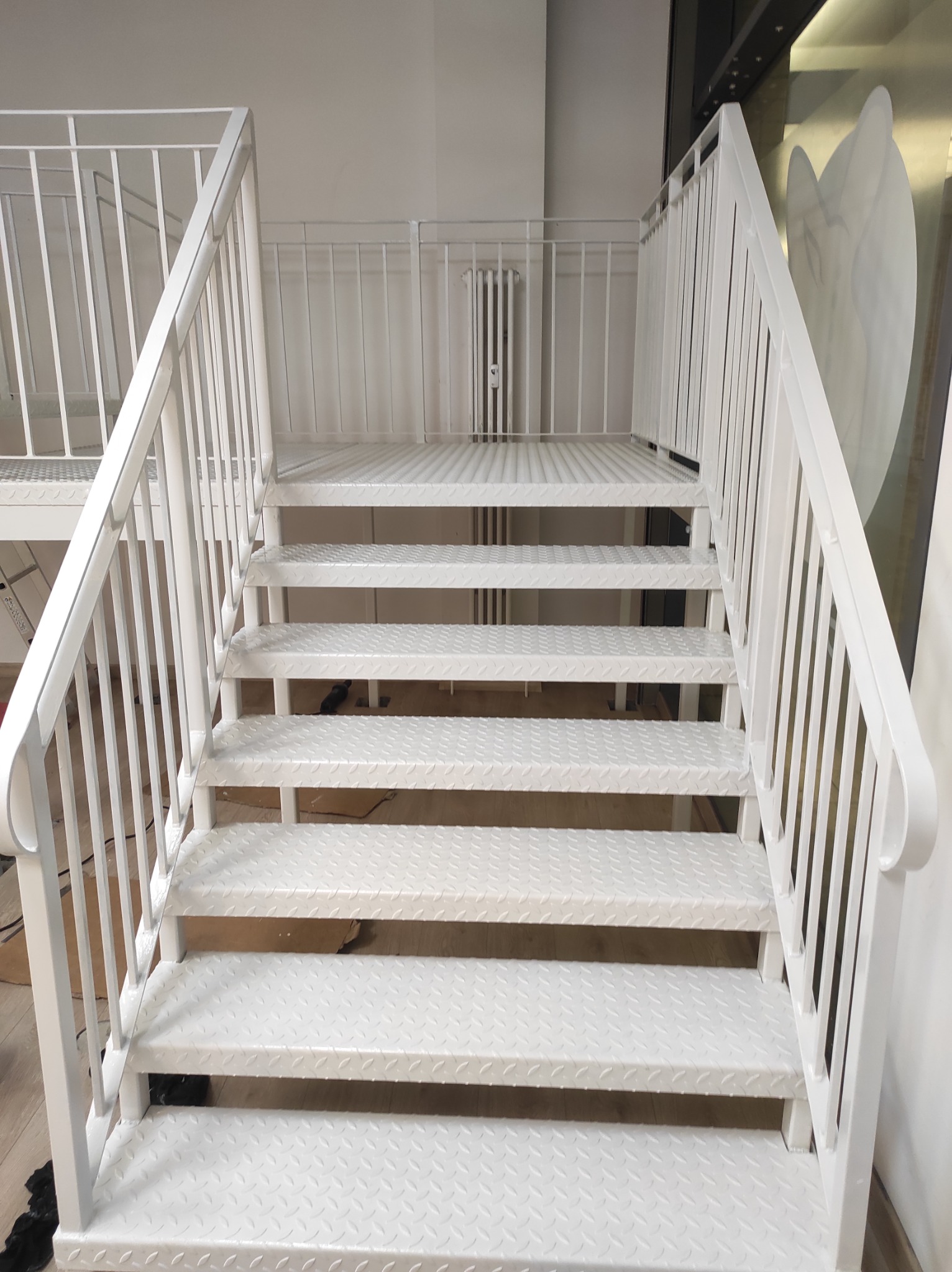 Białe, metalowe schody z ażurową konstrukcją stopni i balustradą, prowadzące do góry, z widoczną kratką na podeście i fragmentem okna z dekoracją w tle.