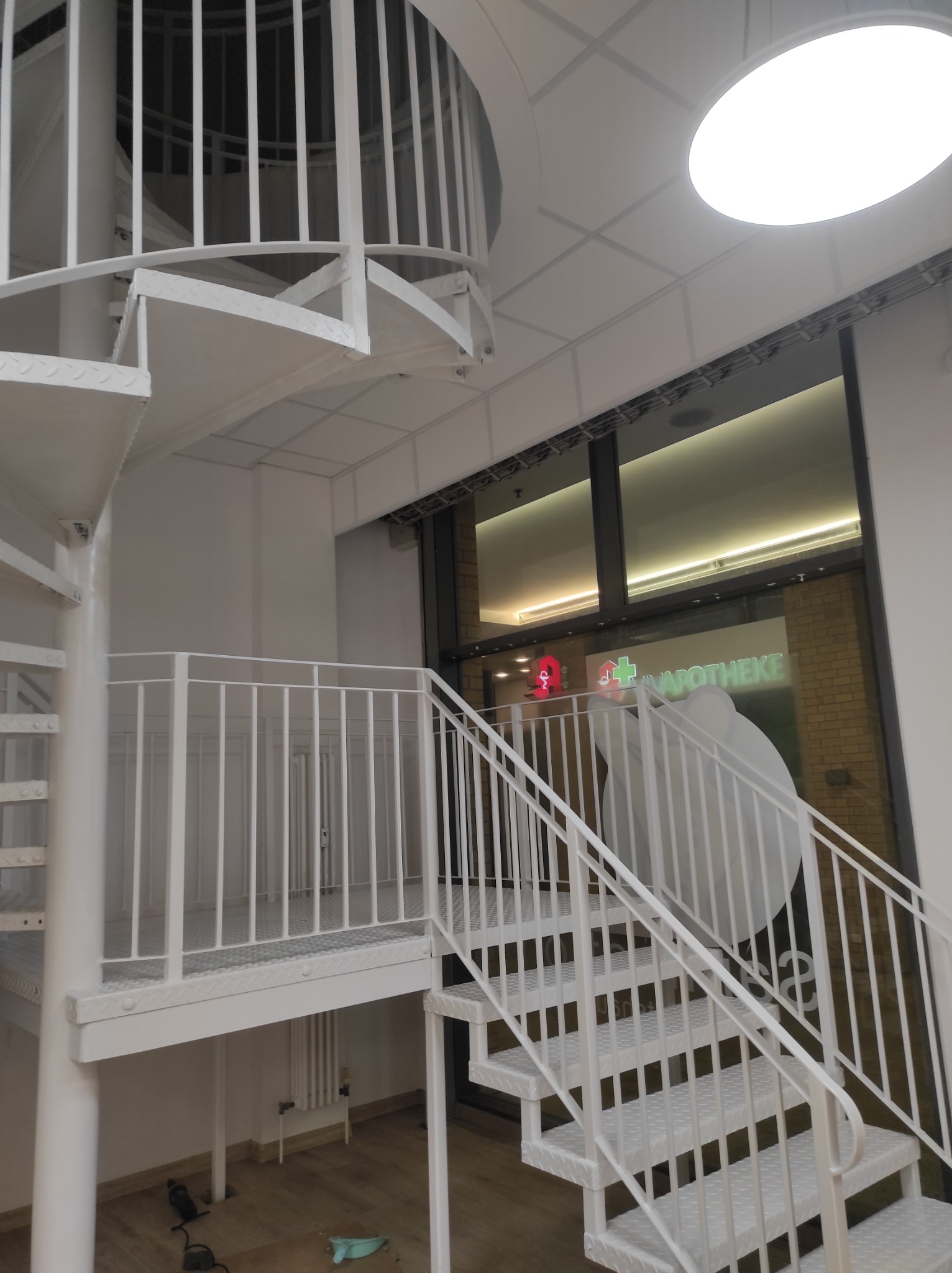 Białe, metalowe schody spiralne z balustradą wewnątrz budynku, widoczne narzędzia na podłodze i okno z neonem apteki w tle.