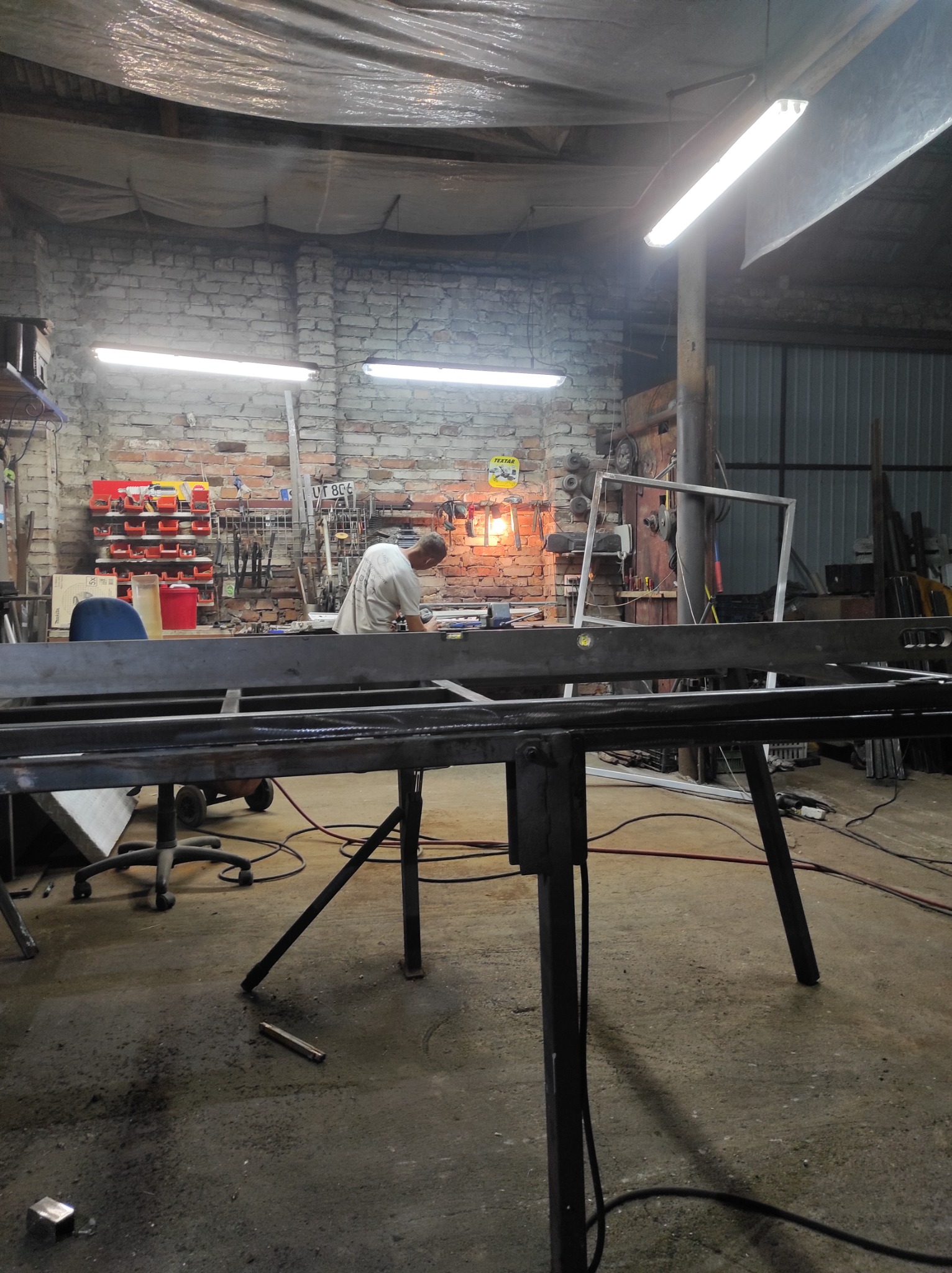 Pracownik w warsztacie przy pracy związanej z obróbką metalu, widoczny spawany element konstrukcyjny na stole warsztatowym, narzędzia i materiały w tle.