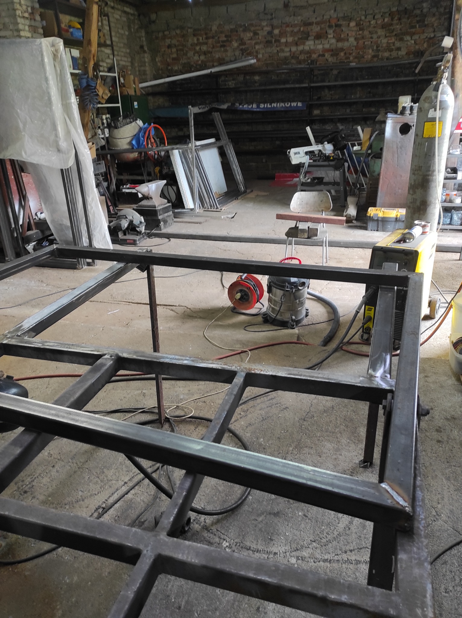 Wnętrze warsztatu z widoczną ramą konstrukcji spawanej z profili stalowych, częściowo zespawana, na tle narzędzi i urządzeń warsztatowych.