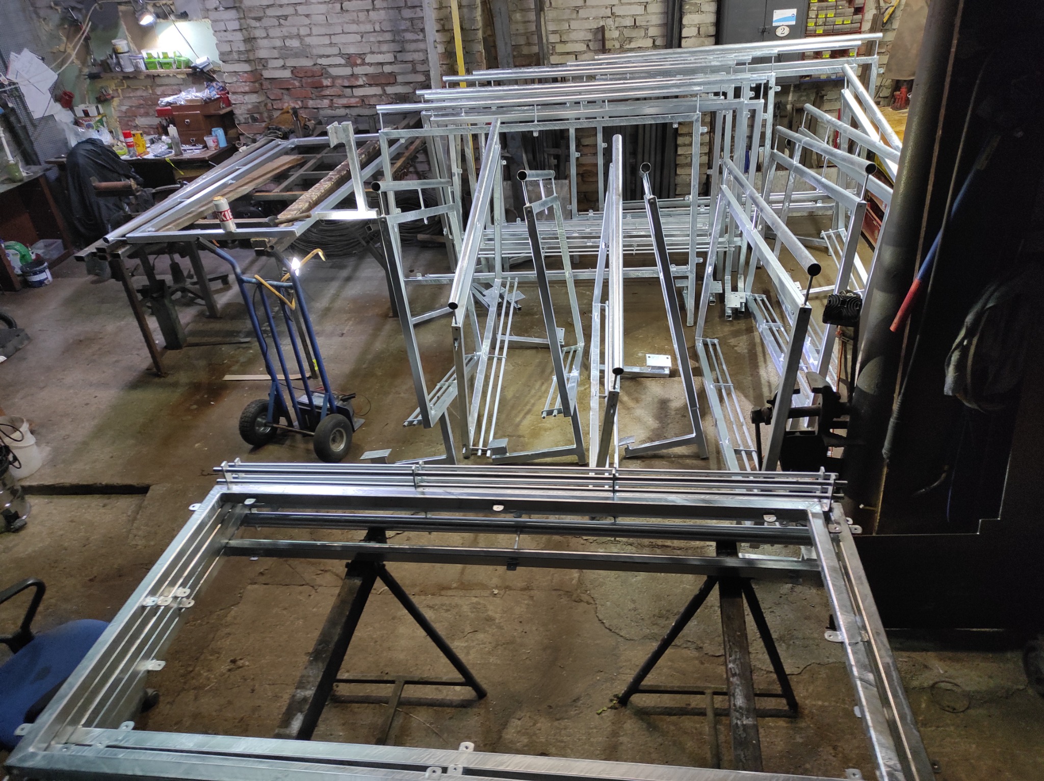 Warsztatowy widok na przygotowane elementy konstrukcyjne z metalu, prawdopodobnie do ogrodzeń lub balustrad, z wózkiem transportowym w tle i widoczną ścianą z cegły.