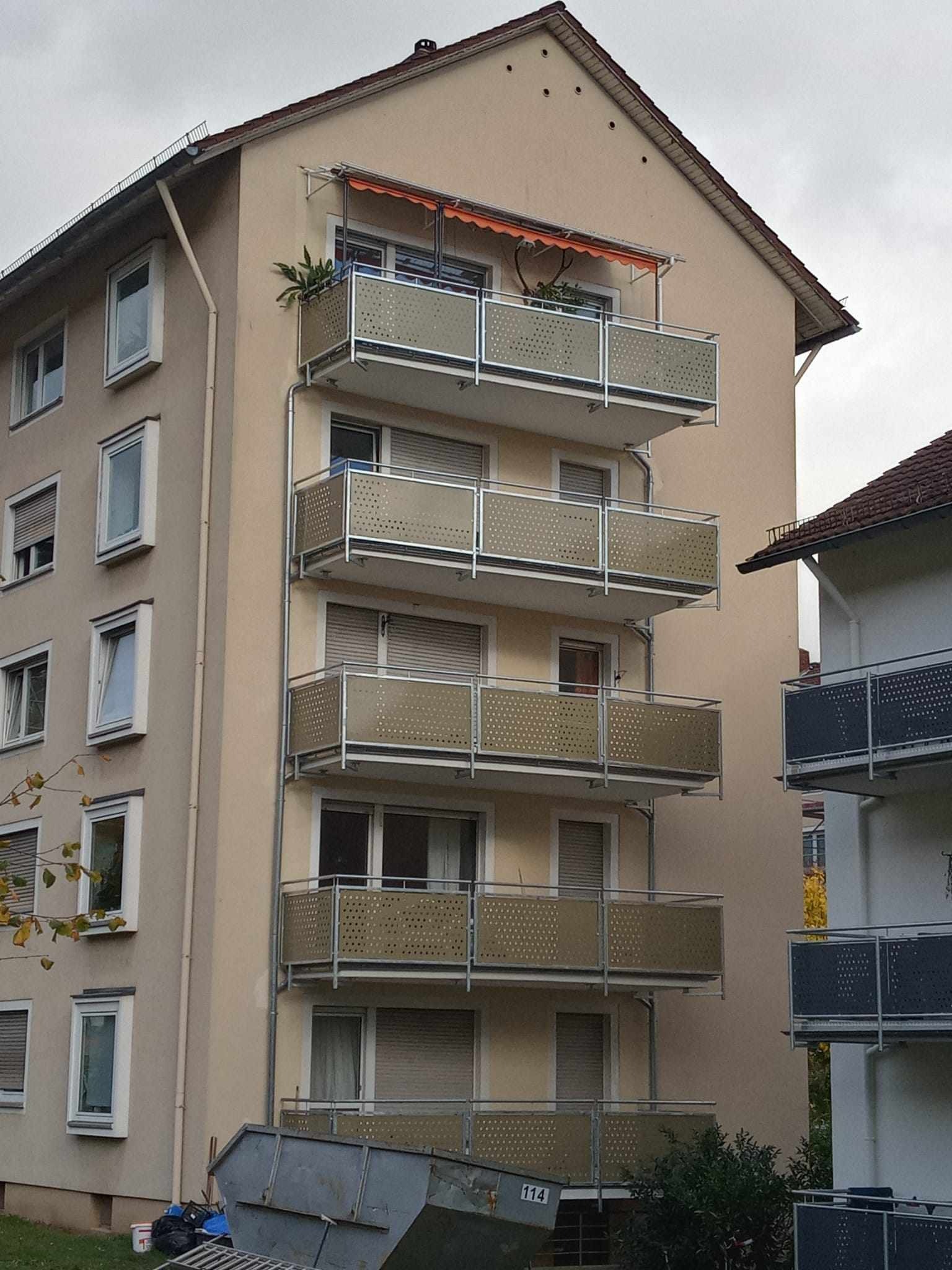 Elewacja budynku wielorodzinnego z balkonami z ażurowymi, metalowymi balustradami w kolorze beżowym i srebrnymi ramami, na jednym z balkonów widoczna roślina w doniczce i pomarańczowa markiza, a u...