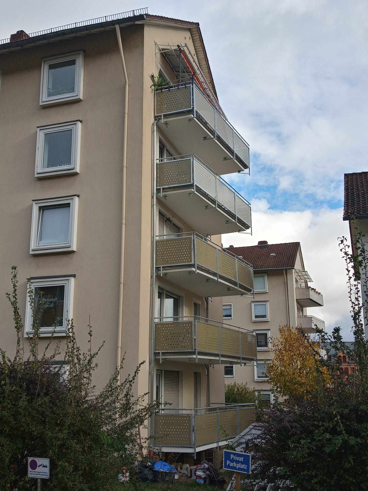 Budynek mieszkalny z balkonami z ażurowymi balustradami w kolorze beżowym i srebrnymi poręczami, widok z dołu na tle pochmurnego nieba.