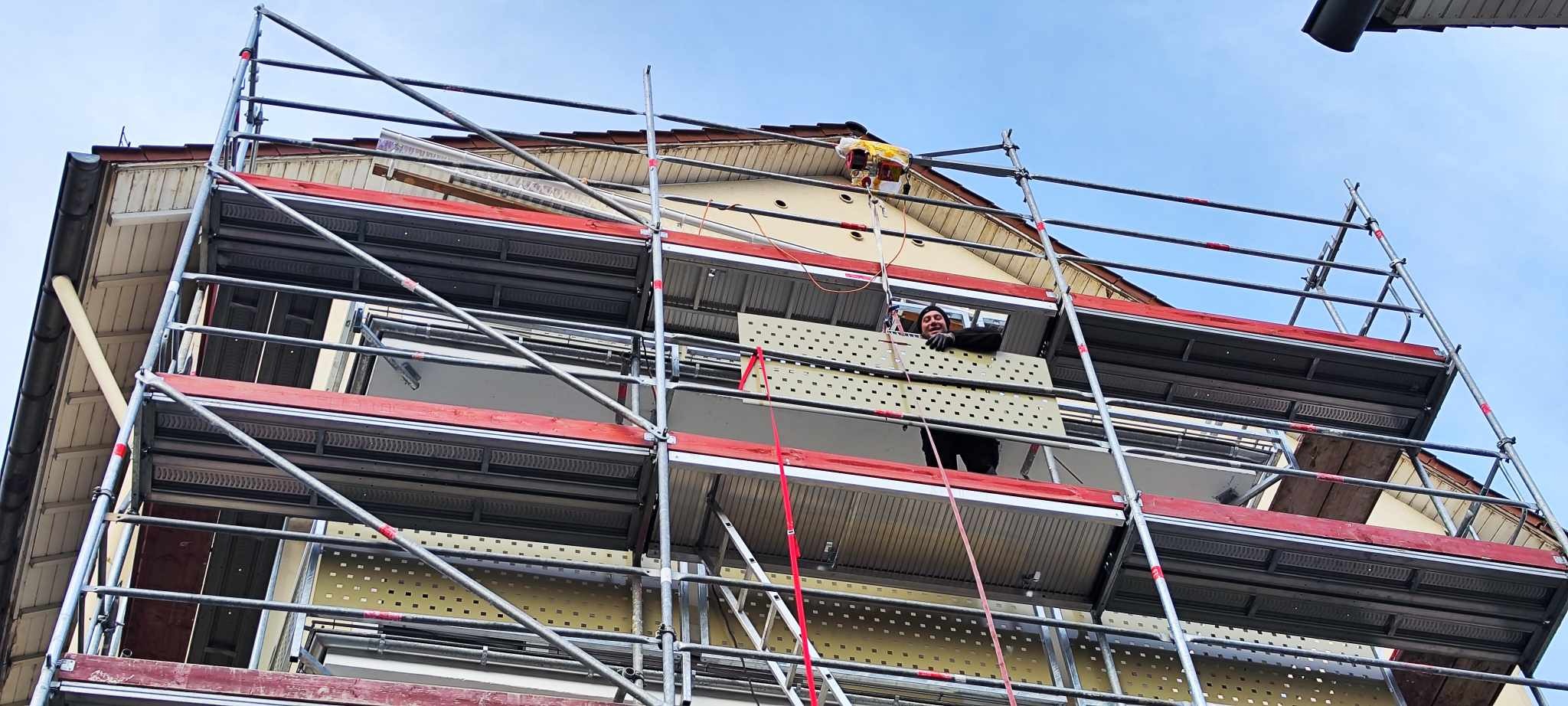 Pracownik na rusztowaniu podczas prac elewacyjnych na dwupiętrowym budynku, widoczny fragment dachu i okna balkonowe.