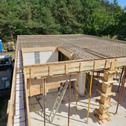 Wieniec stropowy w szalunku drewnianym, zbrojony siatką stalową, wsparty na stemplach, widok z góry na budowę z bloczków betonu komórkowego, w tle las.