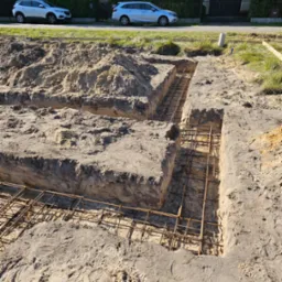 Przygotowanie fundamentów: wykopane rowy z ułożonym zbrojeniem stalowym, widoczne ślady butów na ziemi i dwa samochody osobowe w tle