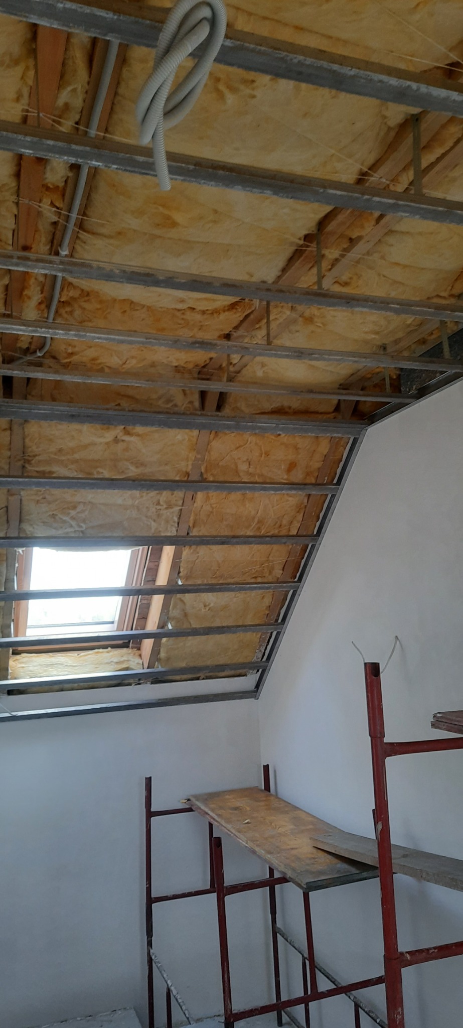 Poddasze w trakcie ocieplania wełną mineralną, widoczne metalowe profile, okno dachowe i fragment rusztowania.