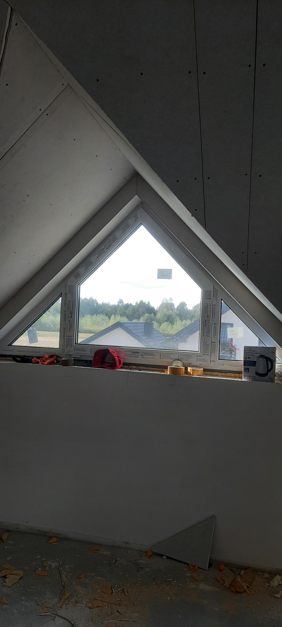 Trójkątne okno dachowe, widok na zewnątrz na domy i drzewa, ściany wykończone płytami gipsowo-kartonowymi, narzędzia i czapka leżą na parapecie.