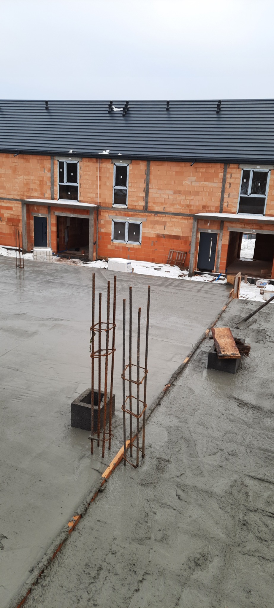 Wylany beton na fundamentach szeregowca z pustaków ceramicznych, zbrojenie pionowe wystaje, widoczne okna wstawione w surowy budynek, pochmurny dzień.