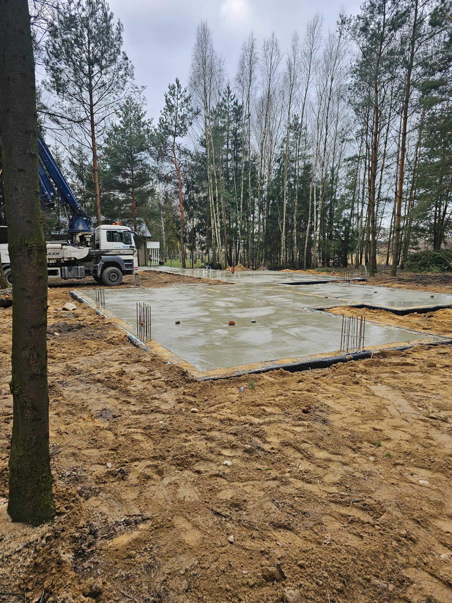 Wylewka fundamentów pod budowę, widoczny betonowy fundament z wystającymi prętami zbrojeniowymi, pompa do betonu w tle, teren budowy otoczony drzewami.